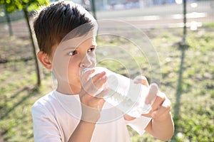 Cute boy sitting on the grass drinks water from a bottle in the summer at sunset. Child quenches thirst on a hot day