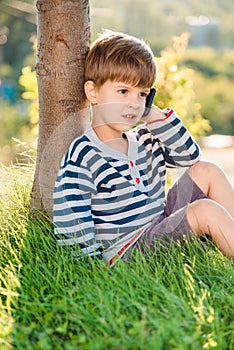 Cute boy sitting on the grass speaks by phone in the summer at sunset. The child communicates on a mobile