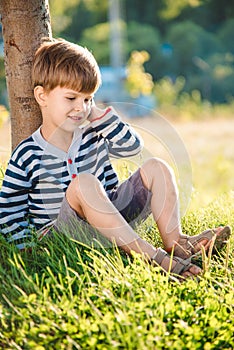 Cute boy sitting on the grass speaks by phone in the summer at sunset. The child communicates on a mobile