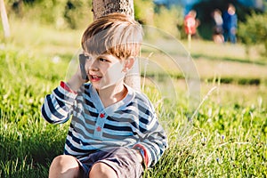 Cute boy sitting on the grass speaks by phone in the summer at sunset. The child communicates on a mobile