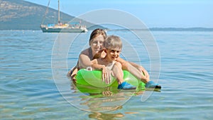 Cute boy sitting in inflatable swimming ring while mother is swimming next to him in the calm sea water at the beach