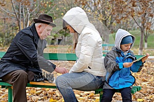 Cute boy sitting on a park bench holding a tablet computer while his mother and grandfather play chess