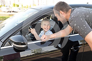 Cute boy is sitting at the wheel in the car, dad is looking out the car window