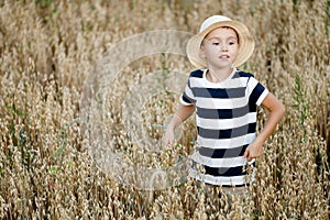 Cute boy in a straw hat and striped T-shirt looks away at a field of oats. He's playing hide and seek