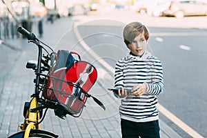 A cute boy in a striped sweater holds a tablet in his hands and stands next to a Bicycle with a backpack hanging from it. The boy