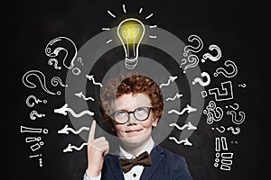 Cute boy student pointing up and looking at lightbulb on blackboard background. Idea concept