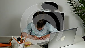 cute boy studying online at his desk. writes assignments in a note and looks at the laptop screen