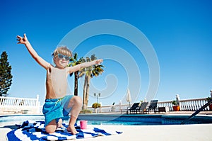 Cute boy in sunshades smiles on towel by pool lifting hands