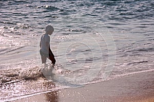 A cute boy walking across a beach next to the ocean at sunset.