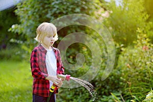 Cute boy watering plants and playing with garden hose with sprinkler in sunny backyard. Funny child having fun with spray of water