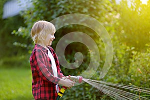 Cute boy watering plants and playing with garden hose with sprinkler in sunny backyard. Preschooler child having fun with spray of