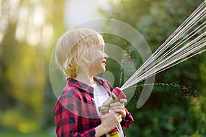 Cute boy watering plants and playing with garden hose with sprinkler in sunny backyard. Preschooler child having fun with spray of