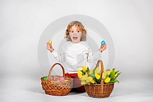 Cute boy with wavy hair sits on a white background next to a basket with Easter eggs