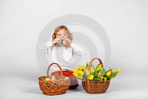 Cute boy with wavy hair sits on a white background next to a basket with Easter eggs