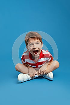 Cute boy wearing red striped shirt, screaming loudly while sitting with crossed legs on studio floor