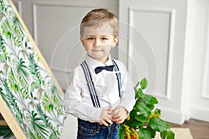 Cute boy in a white shirt and bow tie stands in a room near the tent with a smile.