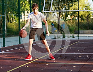 A cute boy in a white t-shirt plays basketball on a city playground. Active teen enjoying an outdoor game with an orange ball.