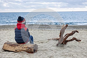 Cute boy in winter jacket and knitted hat siting from behind on deserted beach on log next to snag.