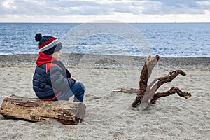 Cute boy in winter jacket and knitted hat siting from behind on deserted beach on log next to snag.