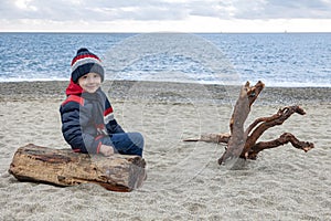 Cute boy in winter jacket and a knitted hat siting on a empty beach on a log next to a snag.