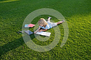 Cute boy writing on notebook laying on grass. Child reading a book in the summer park. Concept of kids learning, study