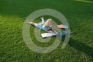 Cute boy writing on notebook laying on grass. Child reading a book in the summer park. Concept of kids learning, study