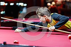 Cute boy in yellow t shirt plays billiard or pool in club. Young Kid learns to play snooker. Boy with billiard cue