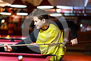 Cute boy in yellow t shirt plays billiard or pool in club. Young Kid learns to play snooker. Boy with billiard cue