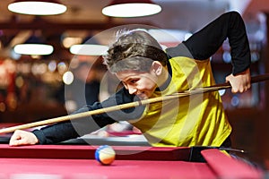 Cute boy in yellow t shirt plays billiard or pool in club. Young Kid learns to play snooker. Boy with billiard cue