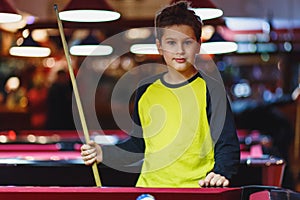 Cute boy in yellow t shirt plays billiard or pool in club. Young Kid learns to play snooker. Boy with billiard cue