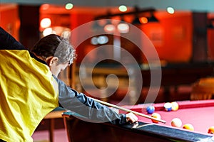 Cute boy in yellow t shirt plays billiard or pool in club. Young Kid learns to play snooker. Boy with billiard cue