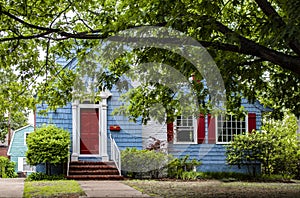 Cute brightly painted blue shingled cottage with red and white trim under a big tree