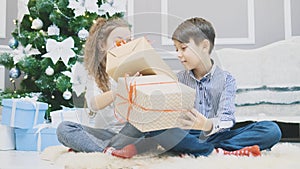 Cute brother and sister sitting in lotos position with Christmas presents in hands, shaking and knocking on it