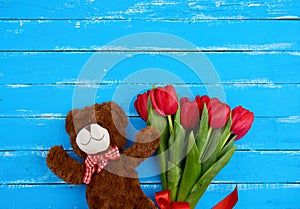 Cute brown teddy bear and bouquet of red blooming tulips with green stems and leaves