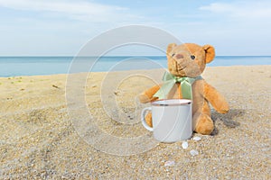Cute brown teddy bear sitting on the beach with a coffee cup. Concept for relaxation, comfort and holiday with copy space