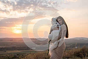 Cute brunette girl in beige dress hugs her samoed puppy, beautiful sunset on the background