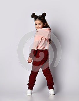 Cute brunette kid girl in modern fashion pink brown hoodie and pants stands back to us looking back over her shoulder