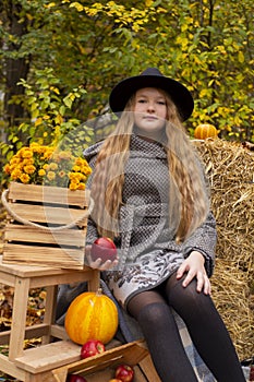 Cute brunette teen girl in black hat and gray coat near autumn elements decoration - pumpkins, apples, plaid, hay. Cosiness,