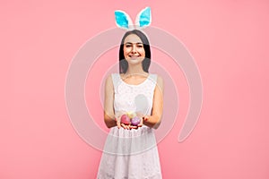 Cute brunette woman in bunny ears holds painted Easter eggs in her hands, Easter, in the studio on a pink background