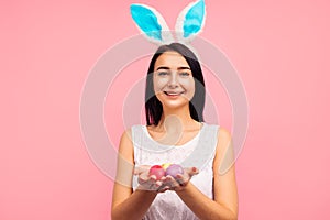 Cute brunette woman in bunny ears holds painted Easter eggs in her hands, Easter, in the studio on a pink background