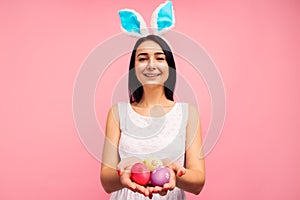 Cute brunette woman in bunny ears holds painted Easter eggs in her hands, Easter, in the studio on a pink background