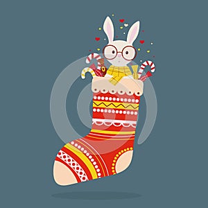 Cute bunny on glasses and with candies sit into christmas sock on blue background