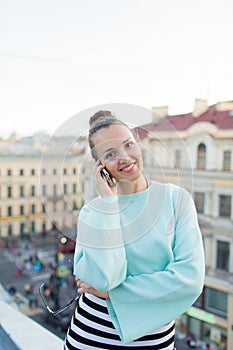 Cute businesswoman in glasses talking on phone and smiling while standing on the roof of the house in the old town