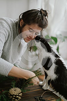 Cute cat helping and kissing young happy woman making rustic christmas wreath. Sweet moments