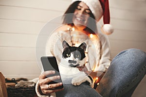 Cute cat looking at phone screen with funny emotions and sitting on happy girl legs in christmas lights. Young hipster woman in