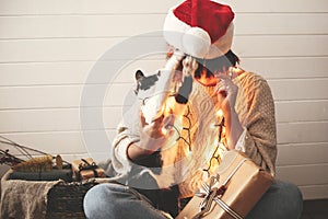 Cute cat playing with santa hat on stylish happy girl in festive christmas lights on background of modern presents. Young hipster