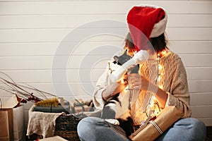 Cute cat playing with santa hat on stylish happy girl in festive christmas lights on background of modern presents. Young hipster