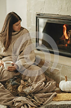 Cute cat and woman with cup of tea sitting together on cozy blanket at fireplace. Adorable tabby kitty relaxing together with