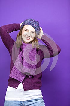 Cute Caucasian Adult Female Woman In Warm Knitted Hat and Violet Scarf with Lifted Hands Pulling Hat Down While Smiling On Purple