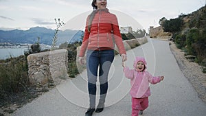 Cute caucasian baby in the hood laughing runs holding mom`s hand trying to chase the camera on coast landscape